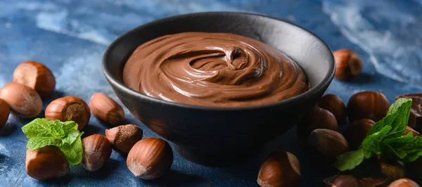 Bowl with tasty chocolate paste and hazelnuts on blue background