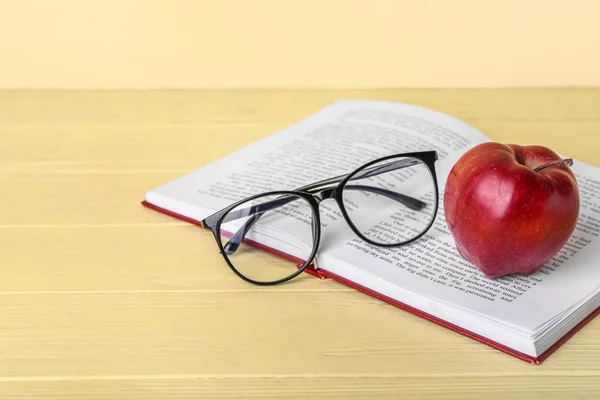 School book with apple and eyeglasses on table against color background