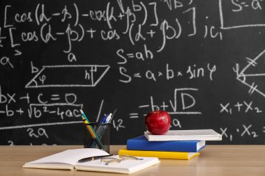 Apple with books, pen cup and eyeglasses on table near blackboard
