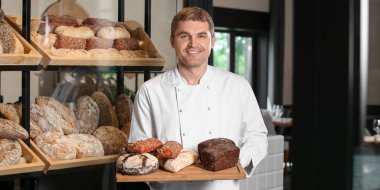 Erkek fırıncı dükkânda taze ekmek çeşitleriyle pano tutuyor.