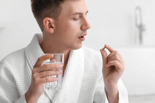 Young man with glass of water taking vitamin supplement in bathroom