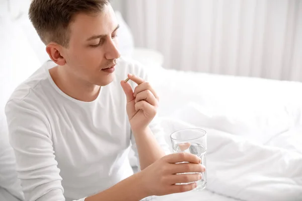 Young man with glass of water taking vitamin supplement in bedroom