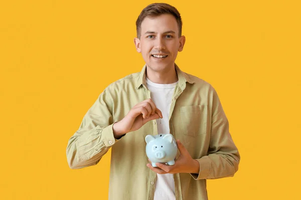 Young Man Putting Coin Piggy Bank Yellow Background — Stock Photo, Image