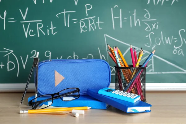 Pencil case with school stationery and eyeglasses on table near chalkboard