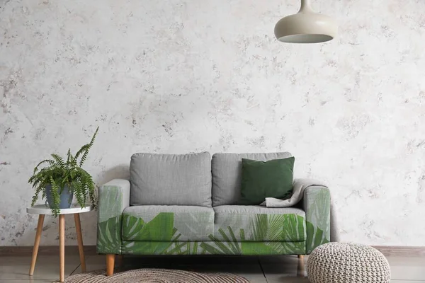 Comfortable sofa with print of tropical leaves and table near light wall in room