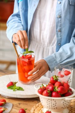 Woman putting fresh mint into glass of cold lemonade in kitchen