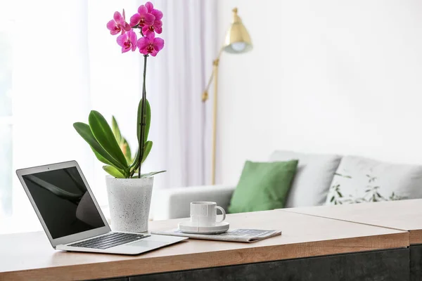 Modern laptop, beautiful orchid flower and cup of coffee on table in light room