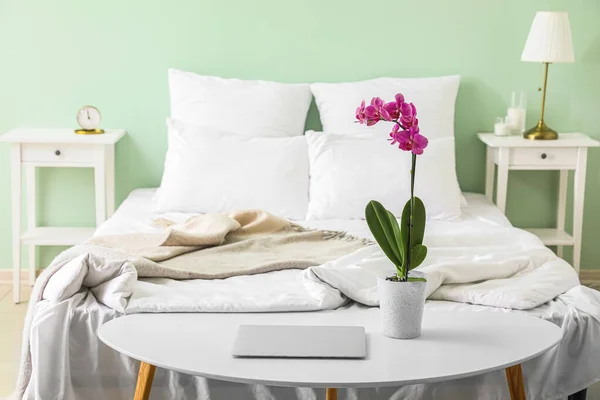 Orchid flower and modern laptop on table near bed