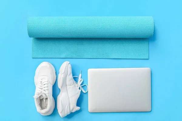 Modern laptop, sports shoes and yoga mat on color background