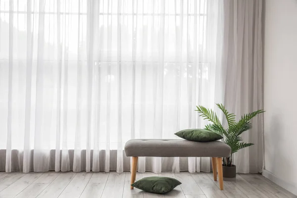 Soft bench with cushions and houseplant near light curtain in living room