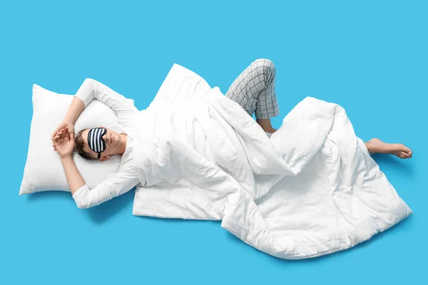 Young man with sleeping mask, pillow and blanket lying on blue background