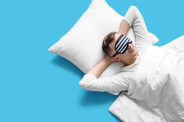 Young man with sleeping mask, pillow and blanket lying on blue background