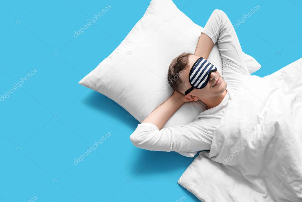 Young man with sleeping mask, pillow and blanket lying on blue background