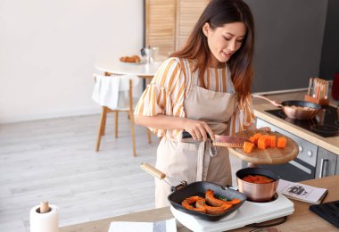 Young Asian woman putting pumpkin into saucepan in kitchen