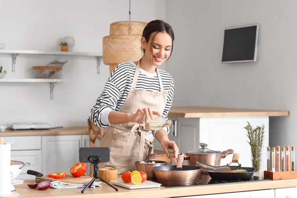 Young woman pouring oil onto frying pan while following cooking video tutorial in kitchen