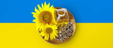 Bottle of oil, seeds and sunflowers in plate on flag of Ukraine, top view