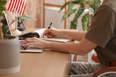 Mature female soldier using laptop and writing in notebook at home
