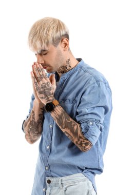 Young tattooed man in denim shirt praying on white background
