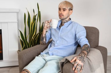 Young tattooed man with cup of coffee sitting in armchair at home