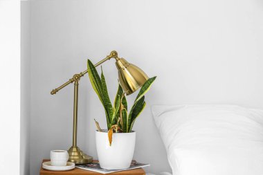 Wilted houseplant, lamp and cup of coffee on table near light wall in bedroom