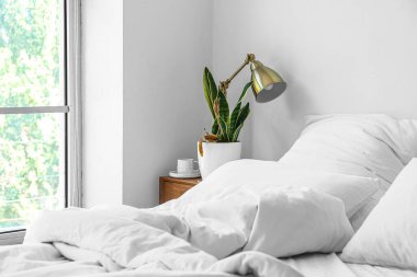 Wilted houseplant, lamp and cup of coffee on table near light wall in bedroom