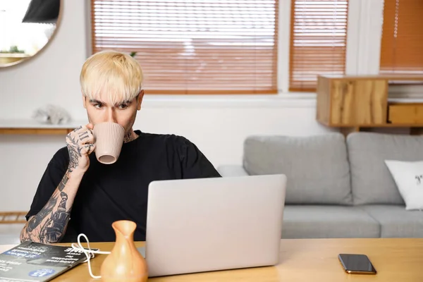 Young tattooed man drinking coffee and using laptop at home