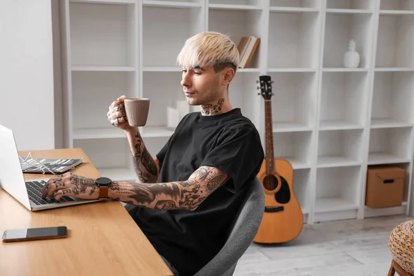 Young tattooed man with cup of coffee using laptop at home
