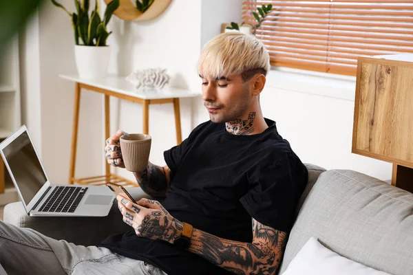 Young tattooed man with cup of coffee and laptop using mobile phone at home