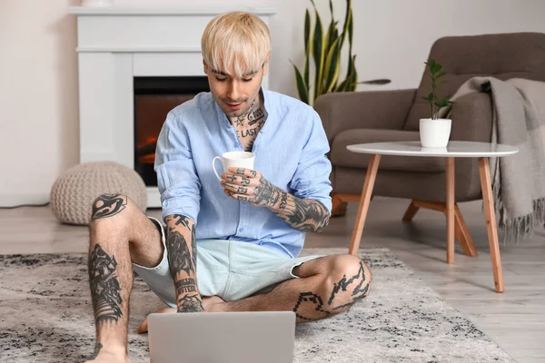 Young tattooed man with cup of coffee using laptop on floor at home