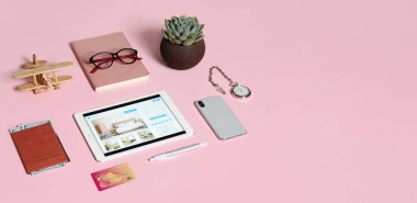 Open page of online booking service on screen of tablet computer and travel accessories on pink background with space for text