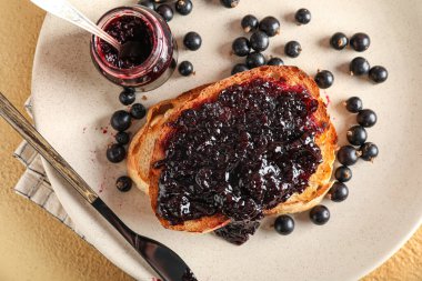 Plate with tasty toast, black currant, jar and knife on beige background, closeup
