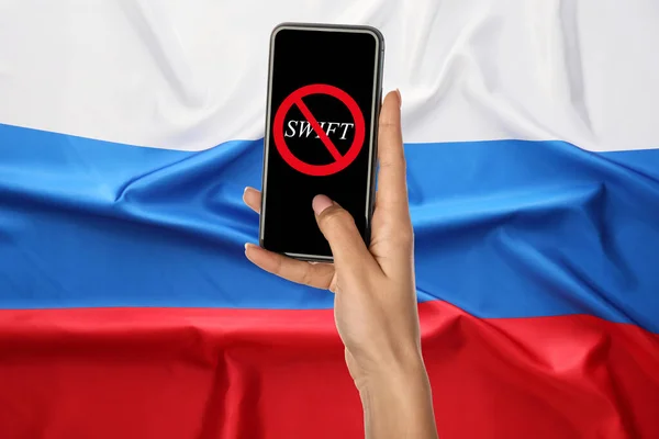 Hand holding smartphone with crossed out emblem of SWIFT on screen against Russian flag. Concept of Ukraine-Russia-related sanctions