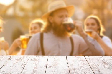 Empty wooden table and young people with beer celebrating Octoberfest outdoors. Mockup for design