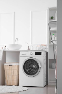 Table with sink, washing machine and basket near light wall in laundry room
