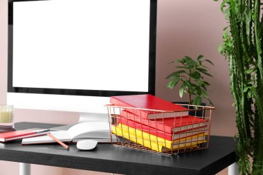 Basket with books, computer and houseplant on table near pink wall