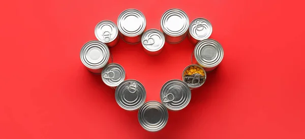 Heart shape made of tin cans with food on red background, top view