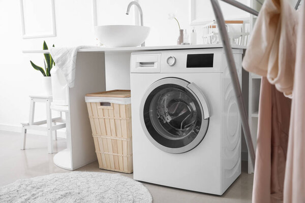 Table with sink, washing machine and basket near light wall in laundry room