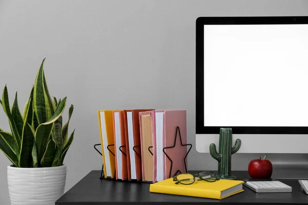 Holder with books, computer, houseplant and apple on table near grey wall