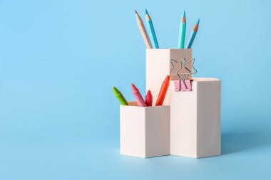 Cups with pencils, crayons and paper clip on blue background