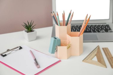Cups with stationery and laptop on table near pink wall, closeup