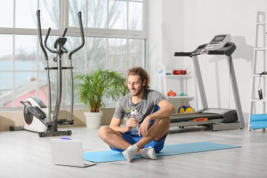 Sporty young man with laptop and bottle of water in gym