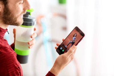 Sporty man with phone and bottle of water at home. Concept of online training
