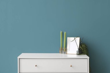 Candles, frame, reed diffuser and houseplant on chest of drawers against blue background