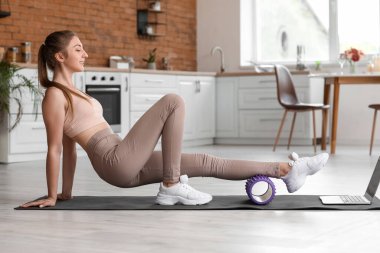Young woman training with foam roller and laptop in kitchen