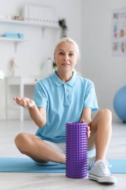 Mature physiotherapist with foam roller sitting on mat in rehabilitation center