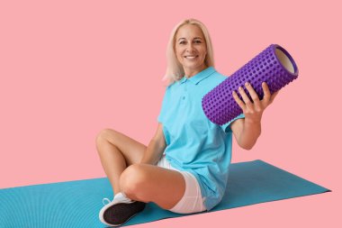 Mature physiotherapist with foam roller sitting on pink background