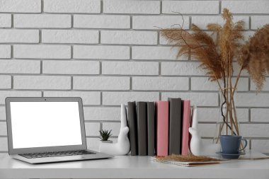 Stylish holder with books, laptop, cup and vase on table near white brick wall