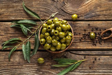 Composition with bowl of green olives, leaves and peppercorns on wooden background