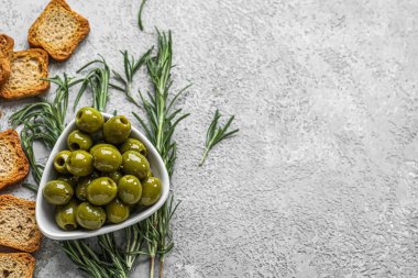 Composition with bowl of tasty green olives, rosemary and crackers on light background