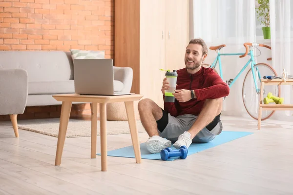 Sporty young man drinking water and using laptop for online training at home